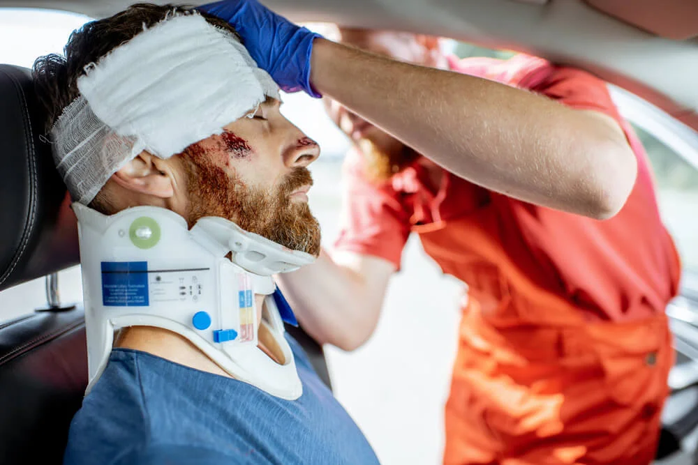Medic applying bandage on the head of victim with serious damages sitting on the driver seat after the road accident