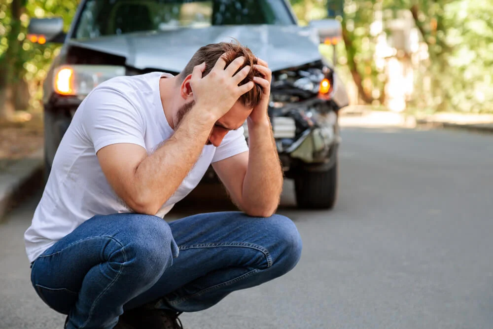 Young white man driver in car accident and holding his head near broken car on the road after a car accident