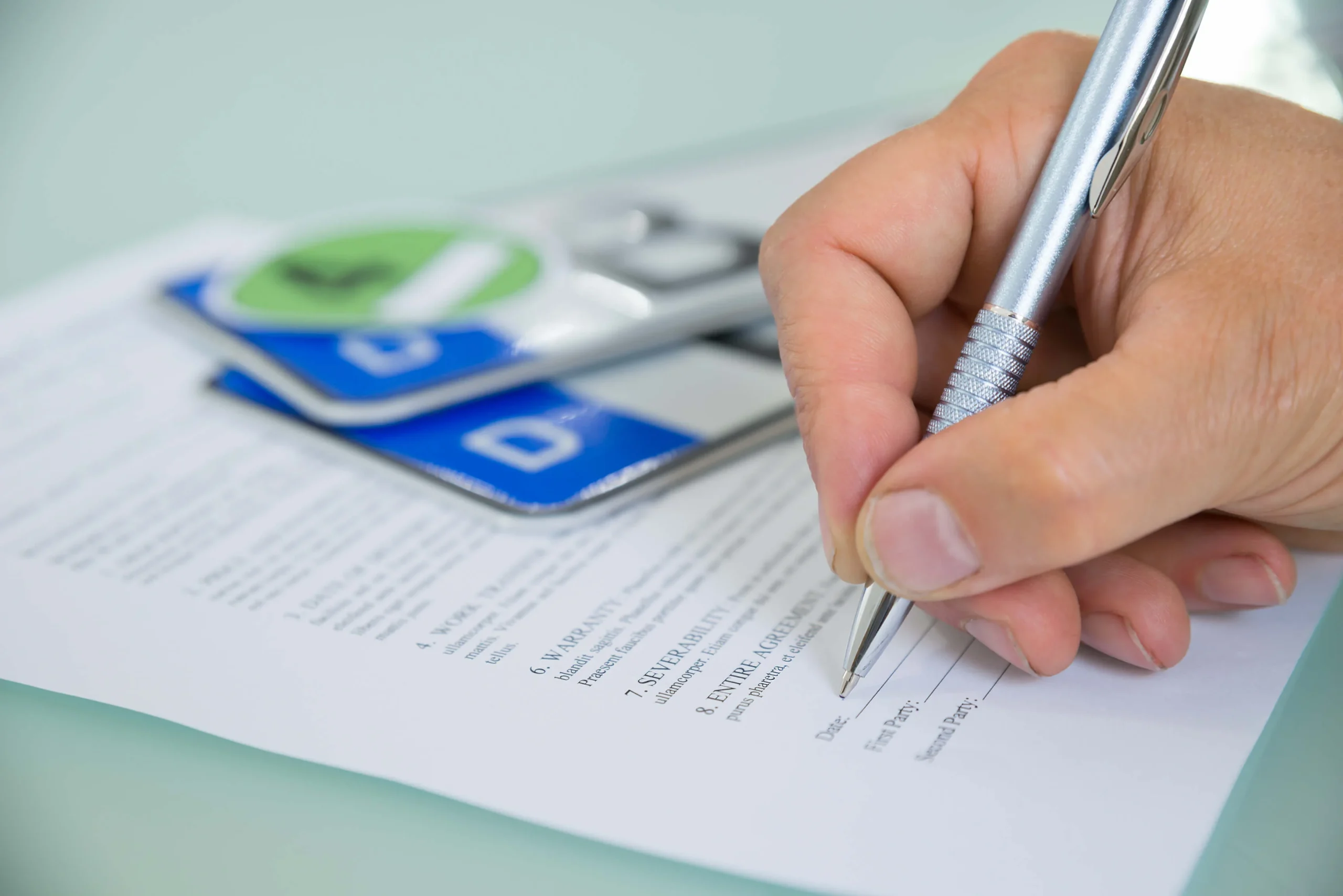 Close-up Of A Hand Filling Car Sale Contract Form With Vehicle Registration Plate On Desk