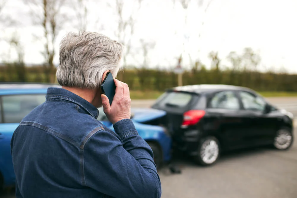 Rear view of senior male driver standing by damaged car after traffic accident reporting incident to insurance company using mobile phone