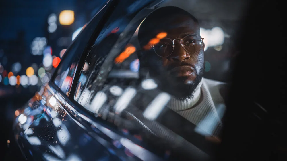 Stylish Black Man in Glasses is Commuting Home in a Backseat of a Taxi on a Rainy Night